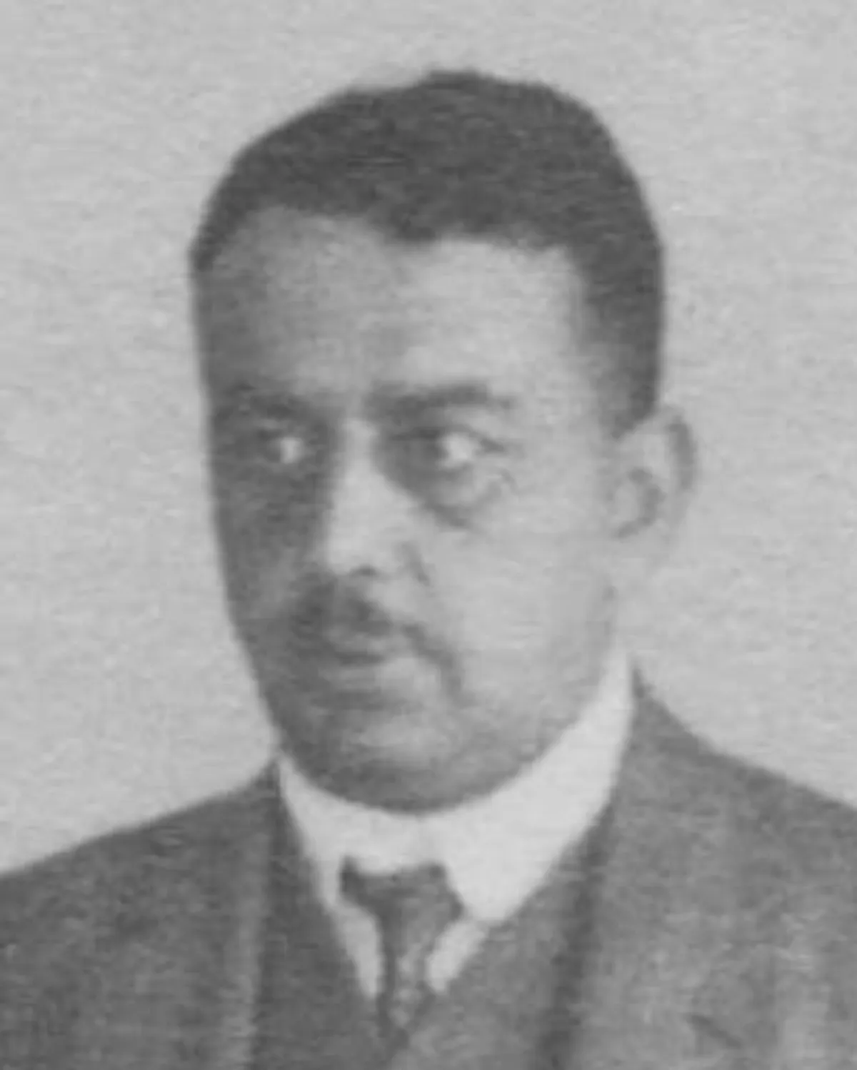 A grainy black and white head-and-shoulders photograph of a man with dark hair and a small moustache, in a suit, white shirt and dark tie.