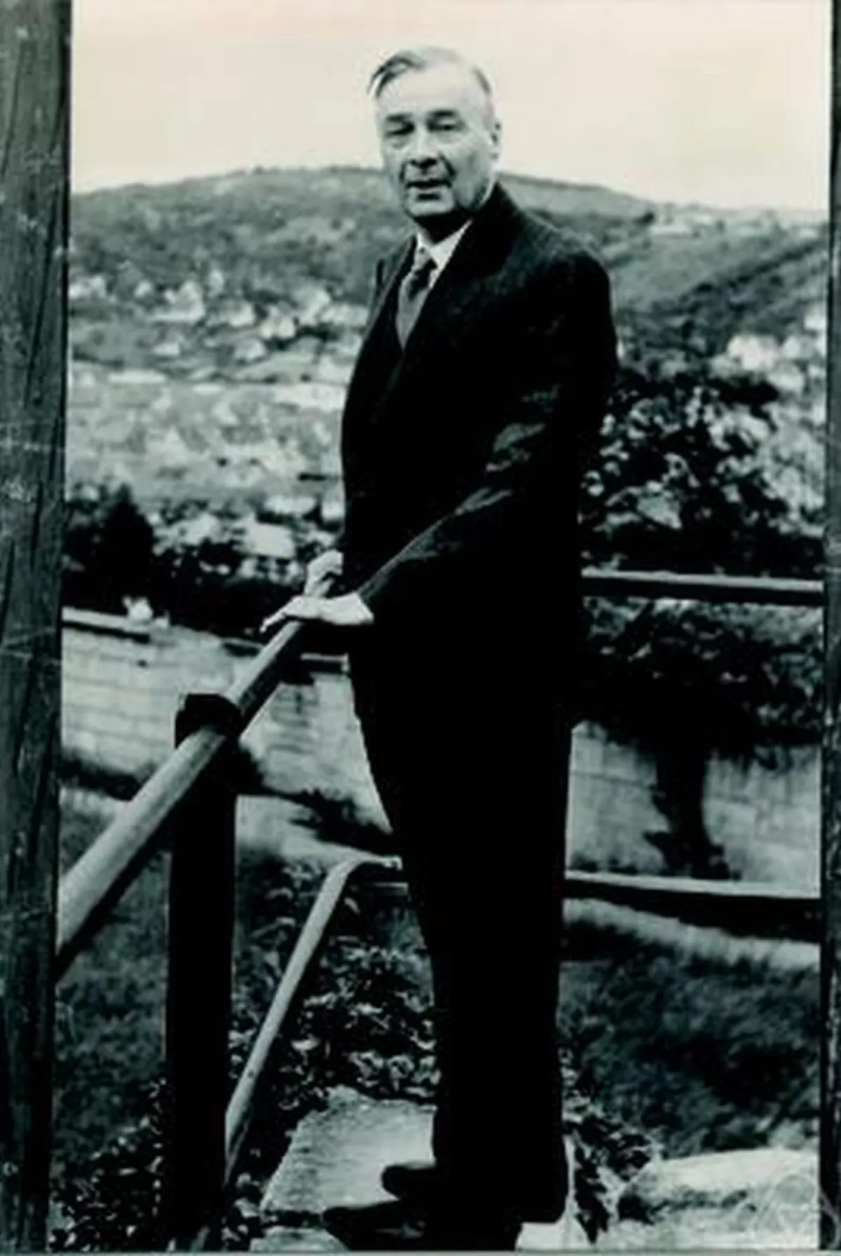 A black and white photograph of a man in a dark suit standing outdoors at a wooden handrail, with a wooded hillside and the roofs of a town behind him.