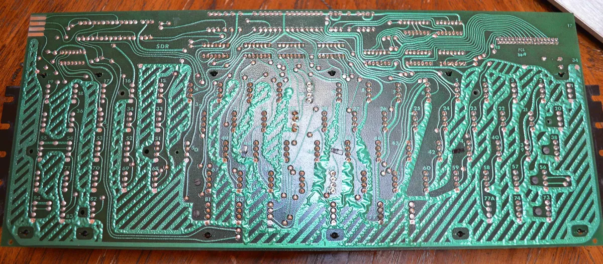 Close-up photograph of the underside of a green circuit board, covered in winding copper traces running between rows of numbered round solder pads.