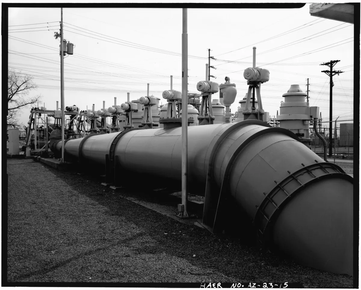 Large-format black and white photograph of a big steel pipe running away from the camera across a gravel yard, with a row of valve assemblies on stands above it and power lines overhead.