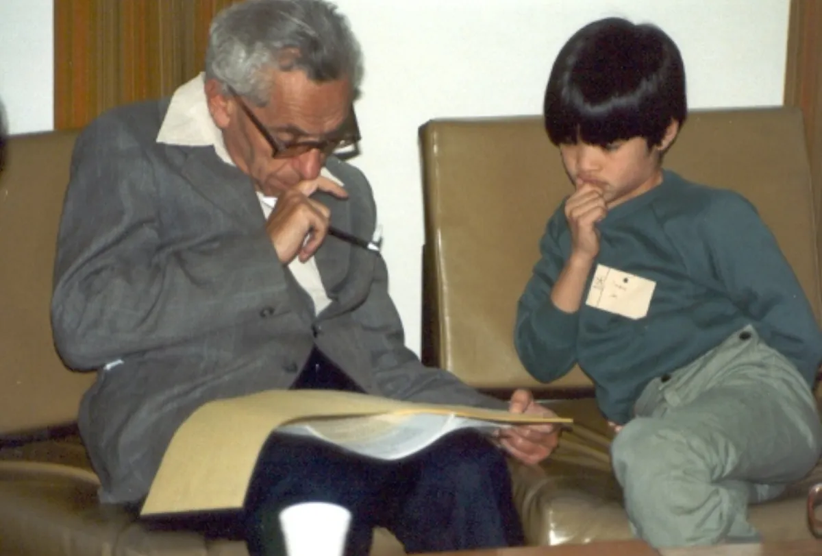 An elderly man in glasses and a young boy wearing a name badge sit on a leather sofa, both looking down at an open folder of papers on the man's lap.