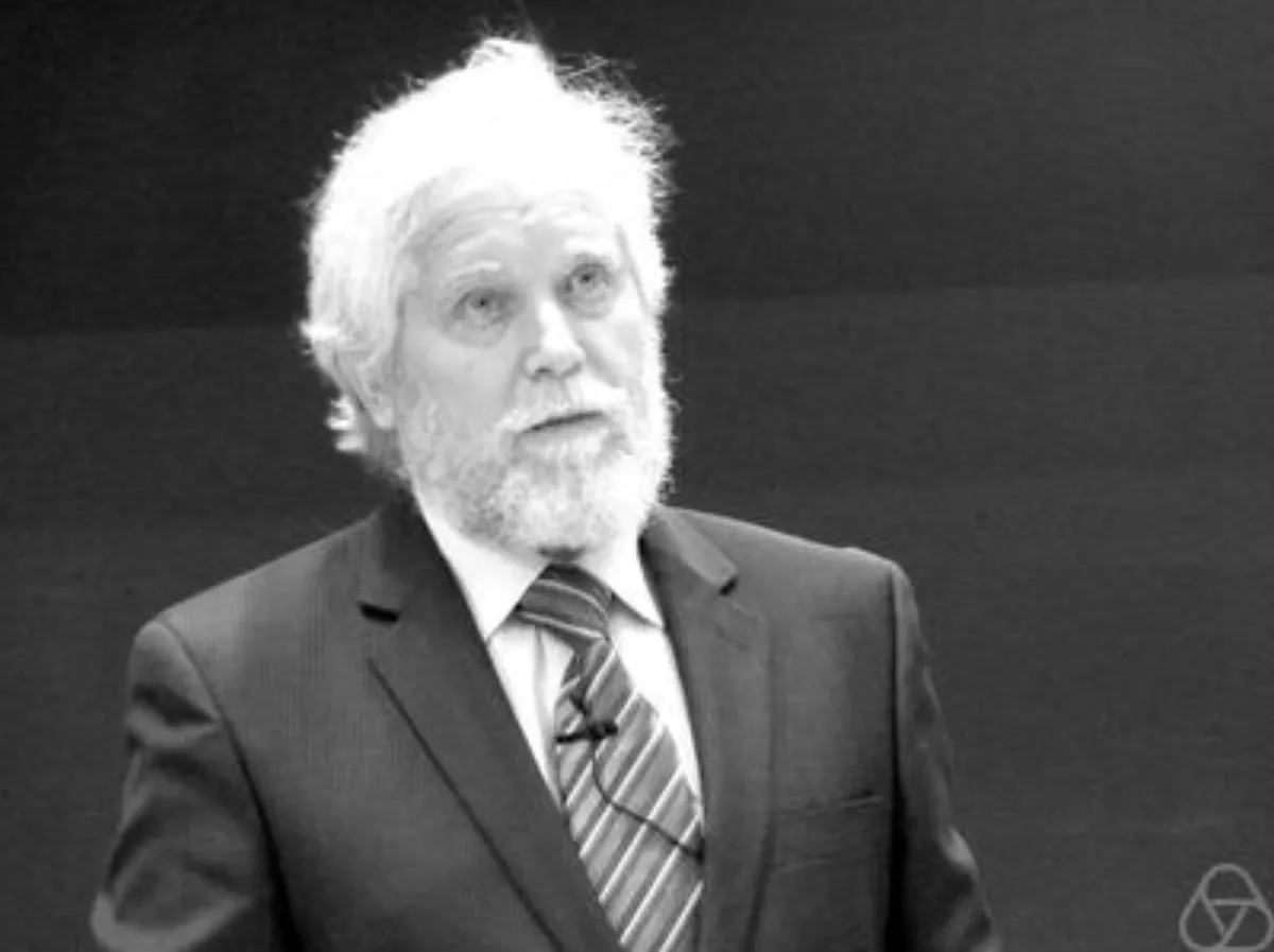 A black and white photograph of an elderly man with white hair and a white beard, in a suit and striped tie with a clip-on microphone, speaking against a dark background.