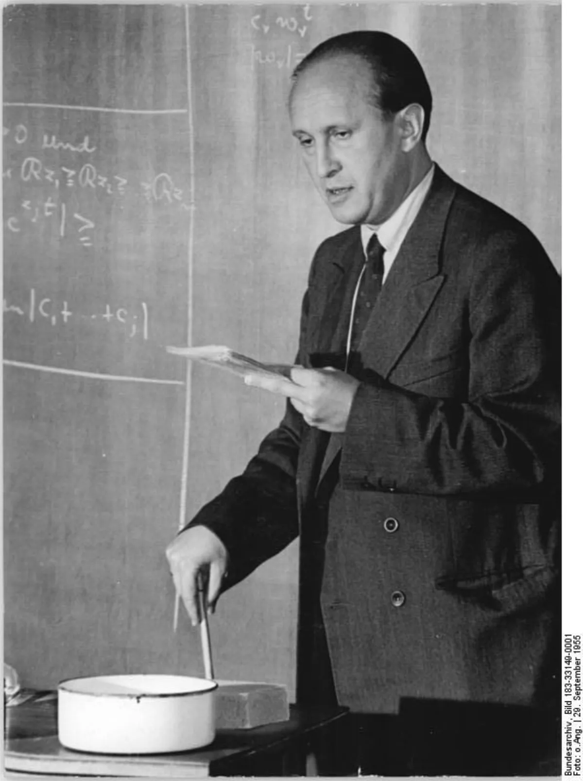 A man in a suit at a blackboard covered in handwritten mathematical formulas, photographed from the side in a lecture room.