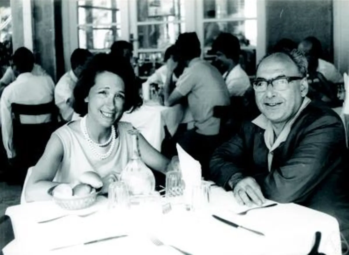 Black-and-white photograph of a man and a woman seated at a restaurant table, both smiling towards the camera.