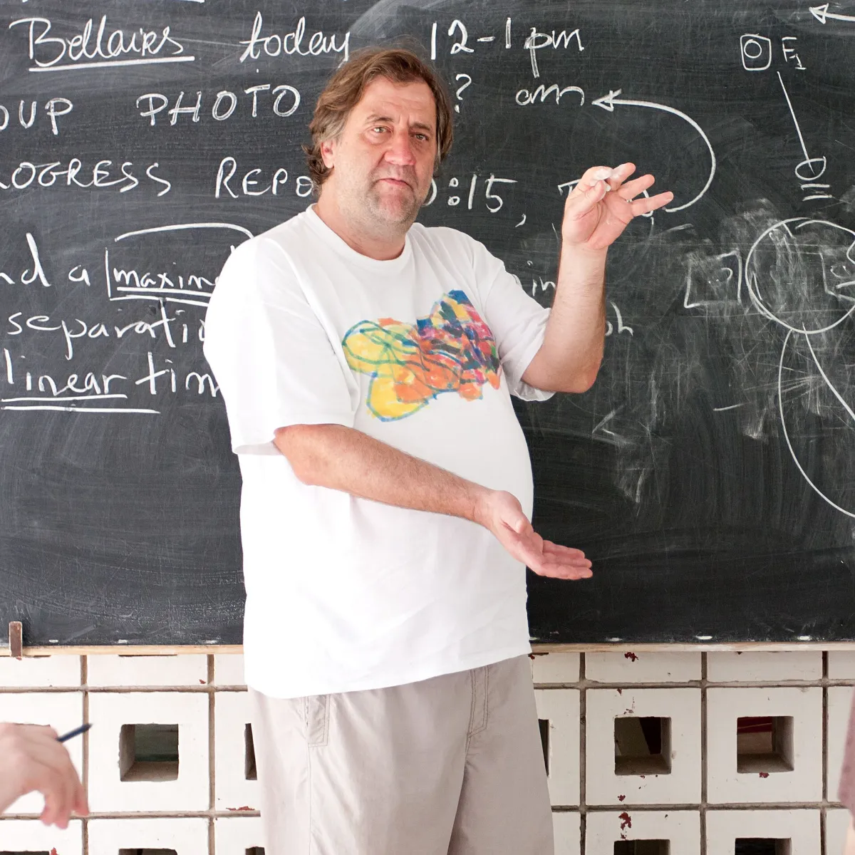 A man gesturing as he speaks in front of a blackboard covered in chalk writing and a handwritten schedule.