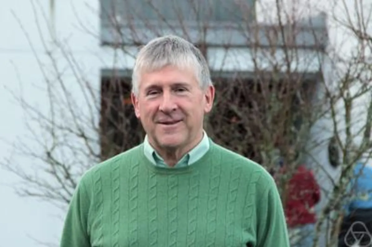 A smiling man with short grey hair in a green cable-knit jumper, photographed outdoors in front of a white building and bare winter trees.
