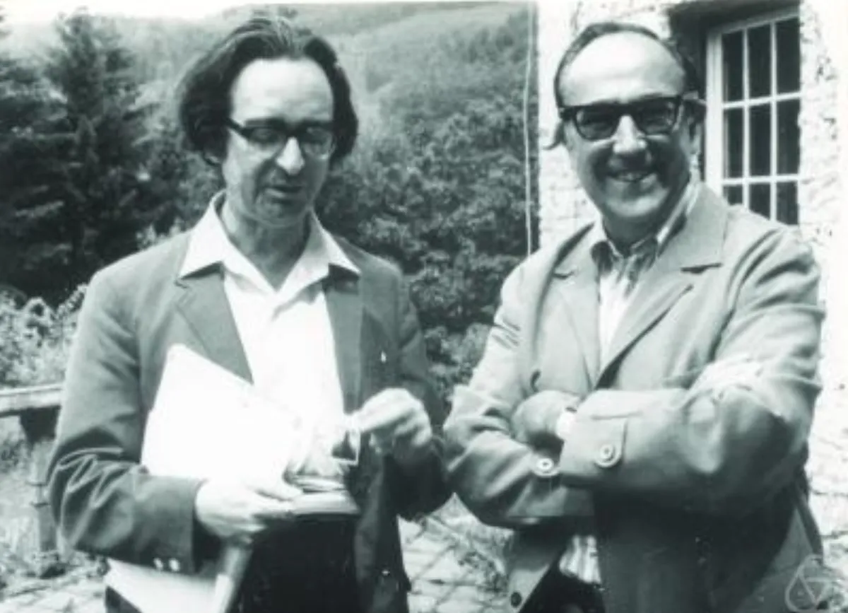 Two men standing outdoors by a stone house, the left one holding papers, the right one in dark sunglasses with his arms folded.