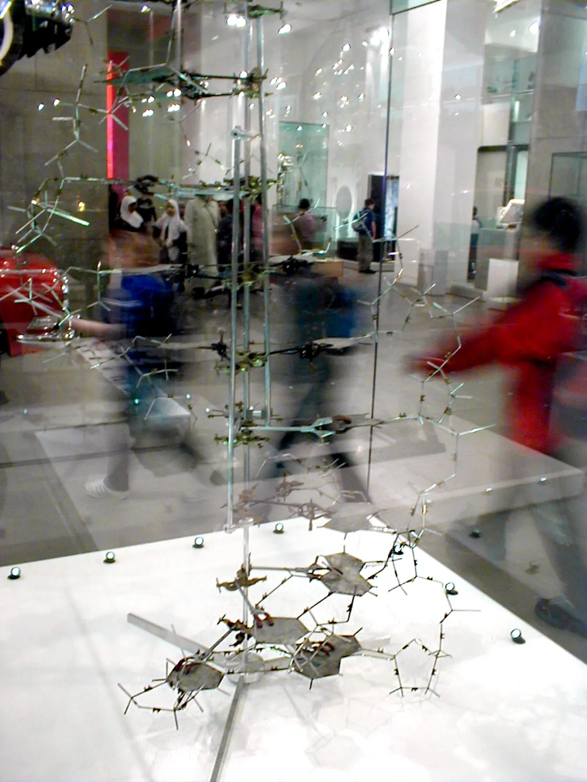 A tall molecular model of metal plates and thin rods on a vertical support, seen through a museum display case with visitors reflected in the glass.