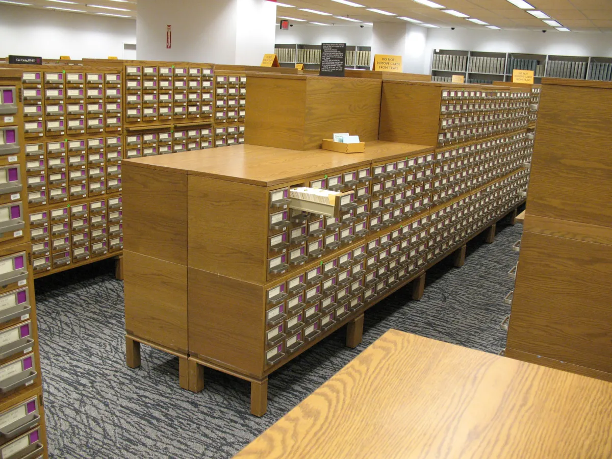 Rows of light oak card-catalogue cabinets fill a carpeted room, one drawer pulled out full of cards, with a wooden counter behind holding a tray.
